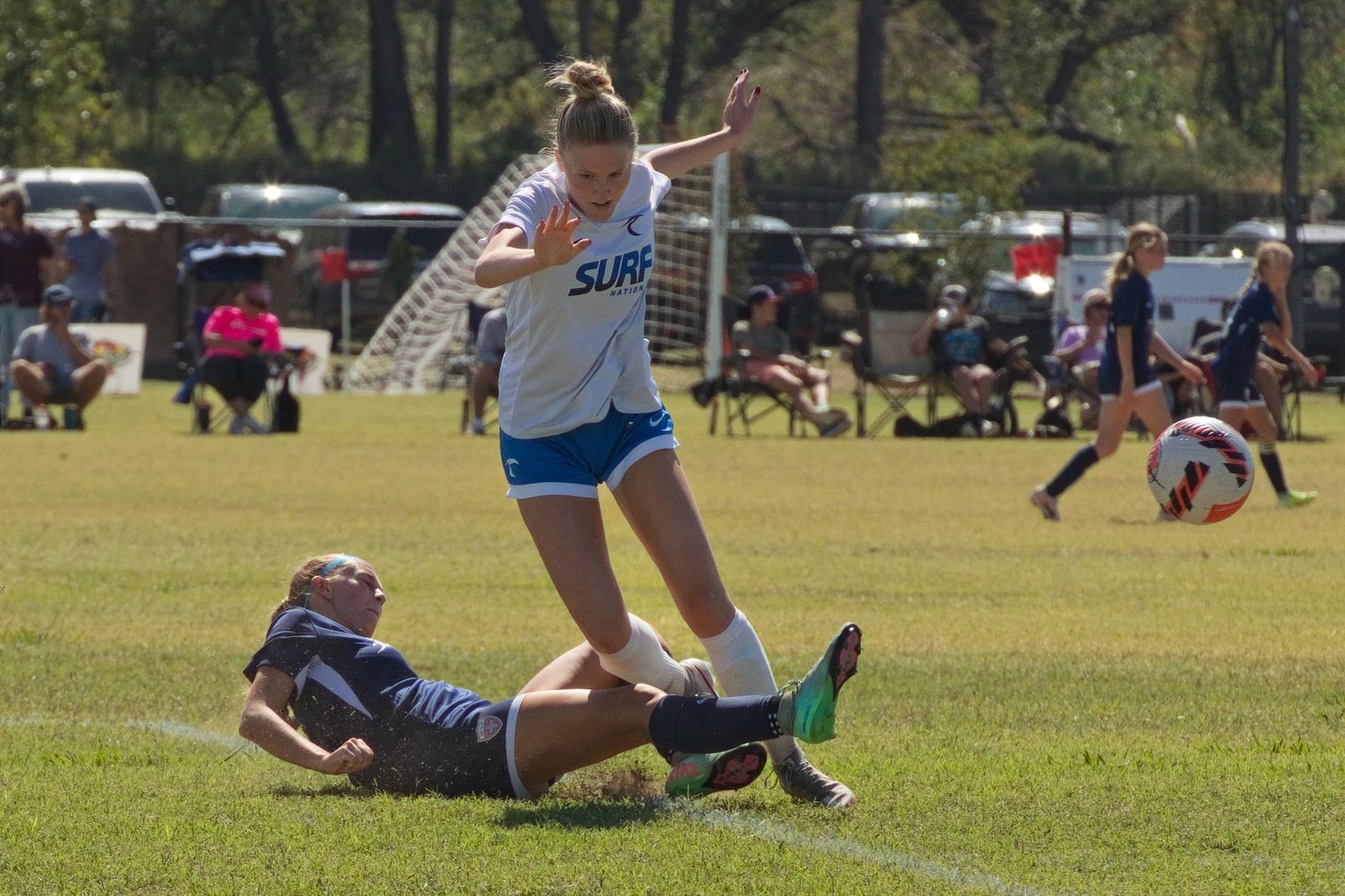 Soccer player hurdling a defender's slide tackle as the ball skips away